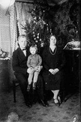Man, kvinna och barn sittande framför julgran i hus.
Nyckelord: Julgran