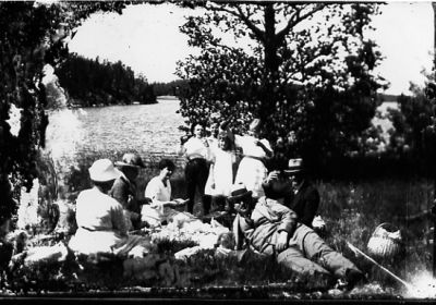 2 män och 3 kvinnor sittande och 4 barn stående i gräset framför vattendrag.
Nyckelord: Picnic