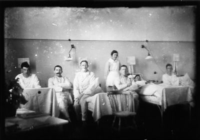 5 män sittande, 1 liggande och kvinna stående i sjuksal.
Nyckelord: Sjuksal sängar