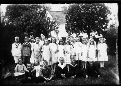 Kvinna och ett antal barn stående eller sittande framför byggnad.
