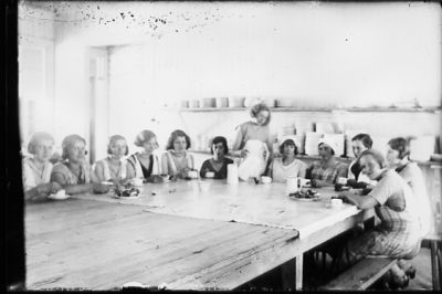 Kvinna stående och 11 sittande i kök.
Nyckelord: Porslin Kaffe