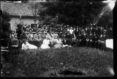 Ett antal personer sittande eller stående framför byggnad.
