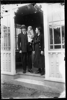 Man med flicka på armen och kvinna stående på veranda.
Nyckelord: Veranda