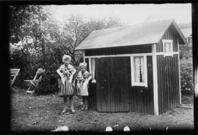 2 flickor i folkdräkt stående framför lekstuga.
Nyckelord: Lekstuga Dräkt