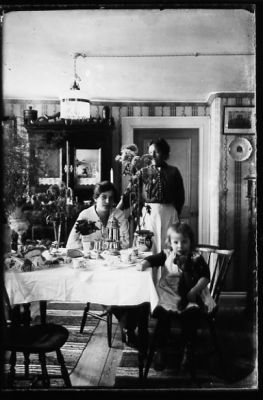 Kvinna stående, kvinna och flicka sittande vid bord i rum.
Nyckelord: Blommor Kaffe Lampa
