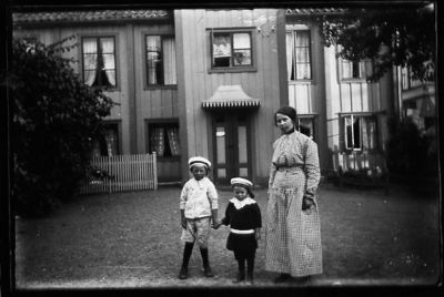 Kvinna och 2 pojkar stående på gård framför byggnad.
