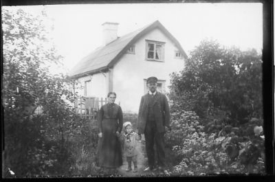 Man, kvinna och ett barn stående i trädgård framför byggnad.
Keywords: Trädgård