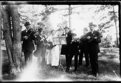 6 män och 1 kvinna stående med musikinstrument i skogsbacke.
Nyckelord: Musikinstrument Notställ