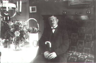 Man sittande i finrum.
Nyckelord: Blommor Lampa Käpp