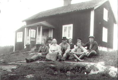 4 män, 2 kvinnor, 1 pojke och 1 flicka sittande framför byggnad.
