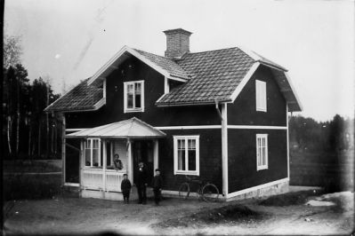 Man, kvinna och 2 pojkar stående framför och på veranda till byggnad.
Nyckelord: Veranda Cykel