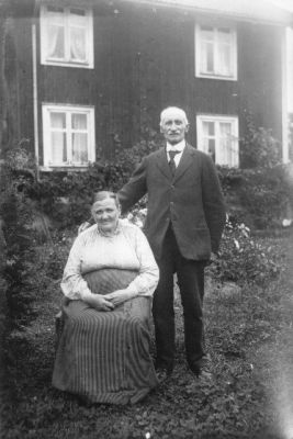 Man stående och kvinna sittande på stol framför byggnad.
Nyckelord: Trädgård Bostad