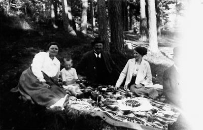2 män, 2 kvinnor och 1 pojke sittande på filt i skogsbacke.
Nyckelord: Filt Picnic