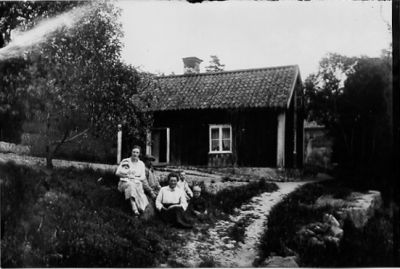 Man, 2 kvinnor och 3 barn sittande på backen framför byggnad.
Nyckelord: Trädgård