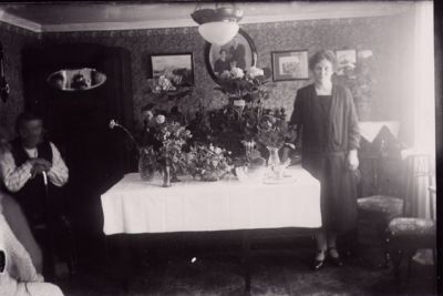 Man sittande och kvinna stående vid bord med blommor, i rum.
Nyckelord: Födelsedag Blommor