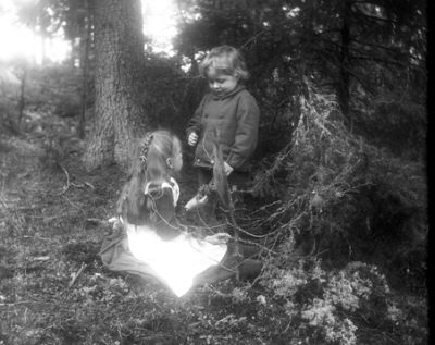 Flicka sittande och pojke stående i skogsmiljö
Nyckelord: Jacka Kjol
