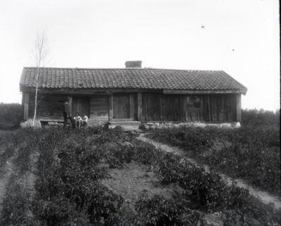 Man och 2 barn framför låg byggnad på landet
