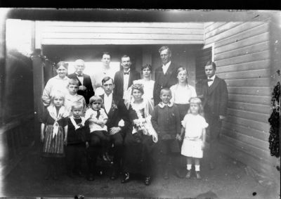 16 personer sittande och stående framför vägg.
Nyckelord: Brudpar Dräkt