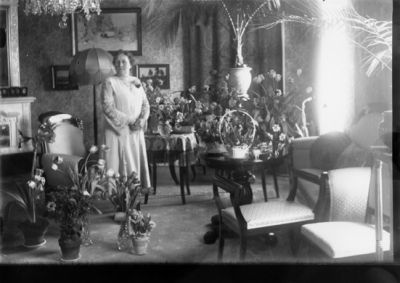 Kvinna stående bland blommor och växter i finrum.
Nyckelord: Möbler