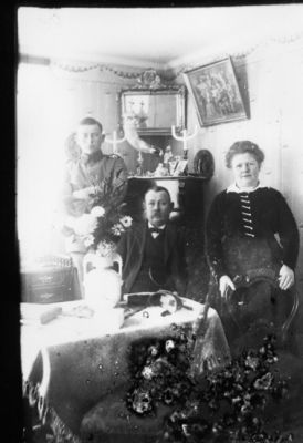 1 man och 1 kvinna stående och 1 man sittande bakom bord i rum.
