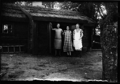 3 kvinnor stående framför stuga.
Nyckelord: Kvinnokläder