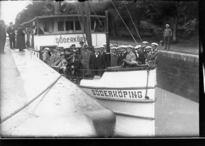 Ett antal stående personer på kanalbåt i sluss.
Nyckelord: Tamp