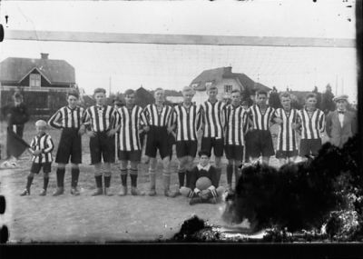 11 stående fotbollsspelare framför fotbollsmål.
Nyckelord: Fotbollslag