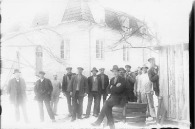 14 män stående och 1 sittande framför byggnad.
