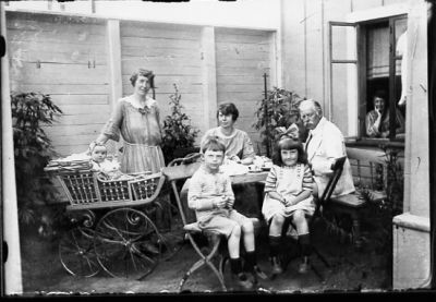 Familj sitter och dricker kaffe på gård.
Nyckelord: Kafferast Barnvagn Dräkt