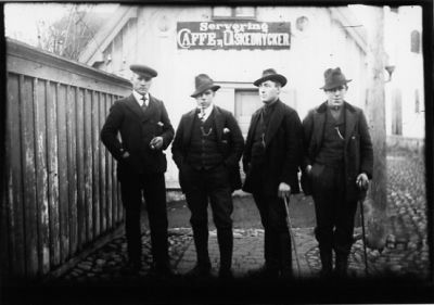 4 män stående framför byggnad.
Nyckelord: Huvudbonad Dräkt Klockkedja Affärsskylt Käpp Kullersten