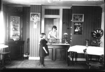 1 flicka stående framför affärsdisk och kvinna stående bakom disken.
Nyckelord: Godis Trattgrammofon