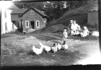 2 kvinnor och 3 barn på gårdsplan tillsammans med några höns.
