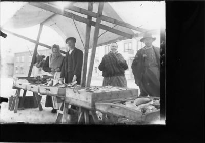 2 män och 2 kvinnor stående bakom fisklådor på torg.
Nyckelord: Torgstånd Fisk