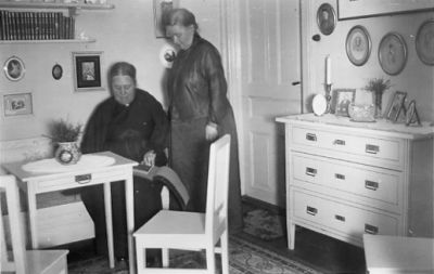 1 sittande och 1 stående kvinna i rum.
Nyckelord: Möbler