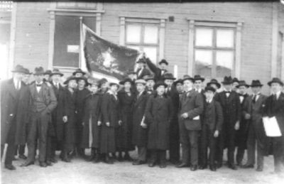 Ett antal personer stående framför byggnad.
Nyckelord: Flagga Byggnad