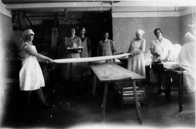 7 stående kvinnor varav 2 drar tvätt.
Nyckelord: Tvättstuga