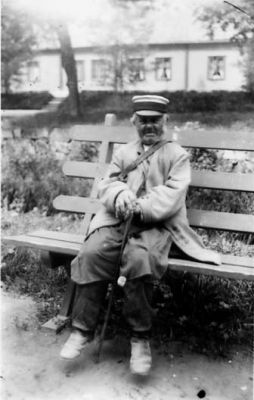 Man sittande på bänk.
Nyckelord: Dräkt Parkbänk Byggnad