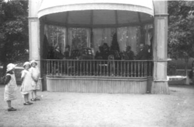 Ett antal personer i musikpaviljong och 3 flickor stående framför.
