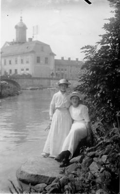 2 kvinnor sittande vid vattendragskant med byggnad i bakgrunden.
