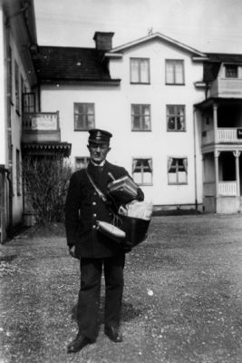 Man i uniform stående med postväska på gård.
