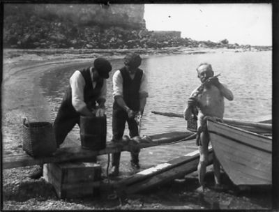 3 män stående och rensande fisk vid strandkant bredvid båt.

