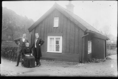 2 stående män och 1 sittande kvinna framför byggnad.
