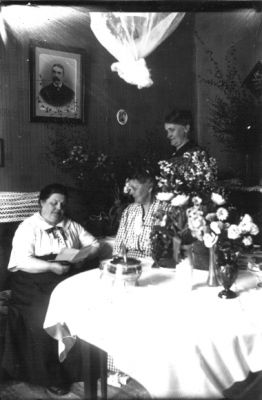 2 kvinnor sittande och kvinna stående vid bord i rum.
Nyckelord: Blommor Tavla