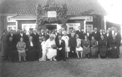 Brudpar med följe stående eller sittande framför byggnad.
