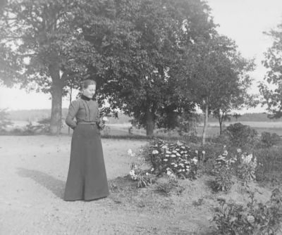Kvinna stående i trädgård med blomma i handen
Nyckelord: Dräkt