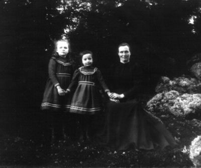 Sittande kvinna med 2 stående flickor i skogsbacke
