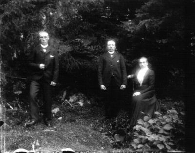 2 stånde män och 1 sittande kvinna i skogsbacke
